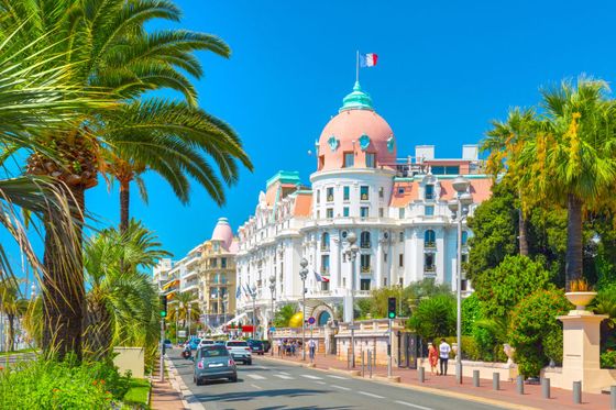 A grand white building with a domed roof and French flag, lined with palm trees on a sunny coastal road, with cars and pedestrians.