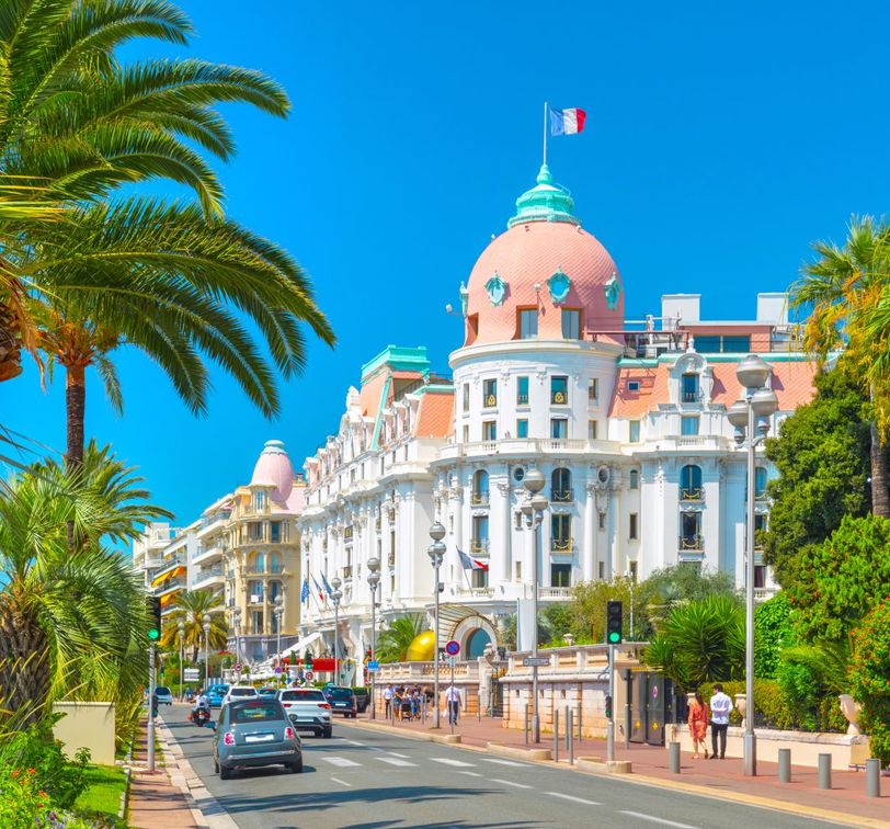 A grand white building with a domed roof and French flag, lined with palm trees on a sunny coastal road, with cars and pedestrians.