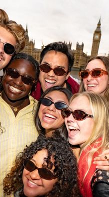 A group of smiling friends wearing sunglasses poses together near Big Ben, with a bridge and the river in the background.