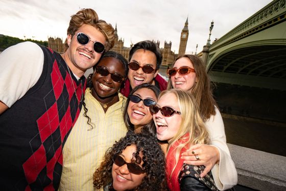 A group of smiling friends wearing sunglasses poses together near Big Ben, with a bridge and the river in the background.