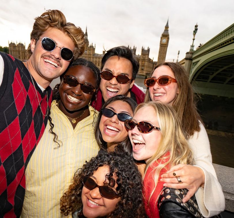 A group of smiling friends wearing sunglasses poses together near Big Ben, with a bridge and the river in the background.