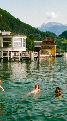 People swimming in a lake near a quaint lakeside hotel with scenic mountains in the background on a sunny day.
