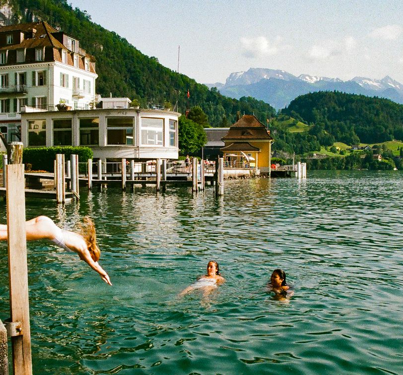 People swimming in a lake near a quaint lakeside hotel with scenic mountains in the background on a sunny day.