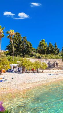 Scenic beach with clear turquoise water, lined with palm trees and a historic church tower, under a bright blue sky.
