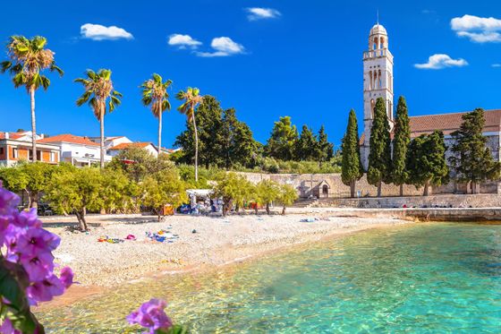 Scenic beach with clear turquoise water, lined with palm trees and a historic church tower, under a bright blue sky.