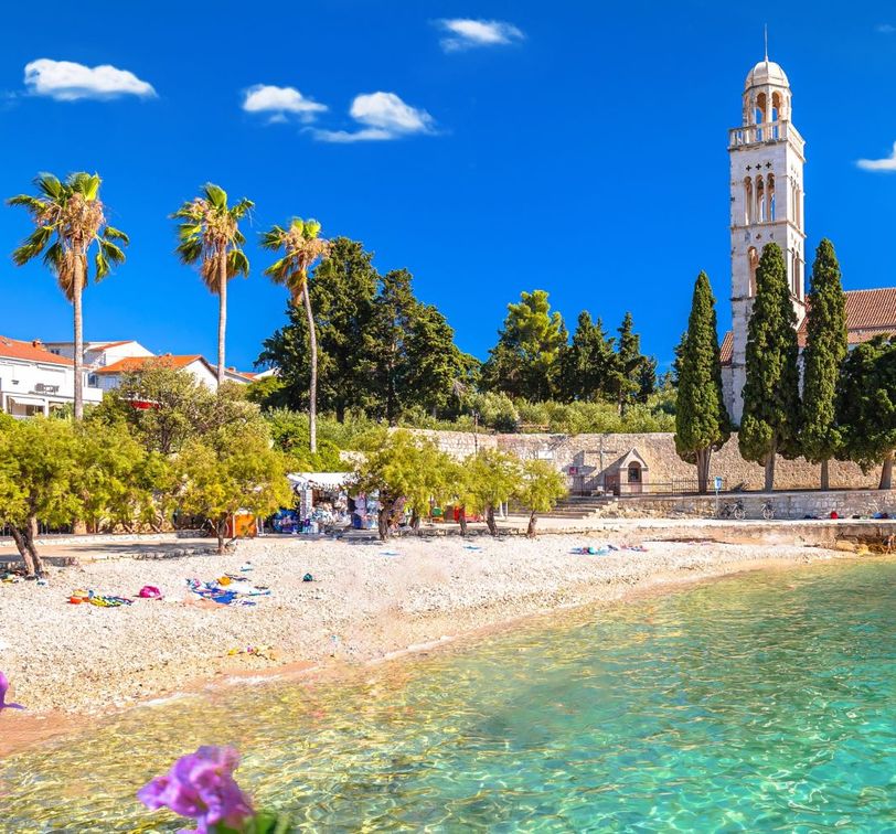 Scenic beach with clear turquoise water, lined with palm trees and a historic church tower, under a bright blue sky.