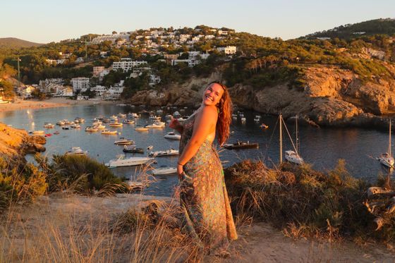 A woman in a sundress smiles on a hilltop overlooking a coastal bay filled with boats, with a cliffside town in the background during sunset.