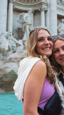 Two smiling women in front of the Trevi Fountain in Rome, with clear turquoise water and detailed marble sculptures in the background.