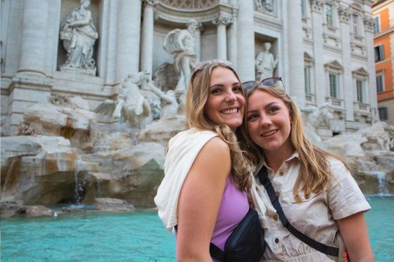 Two smiling women in front of the Trevi Fountain in Rome, with clear turquoise water and detailed marble sculptures in the background.