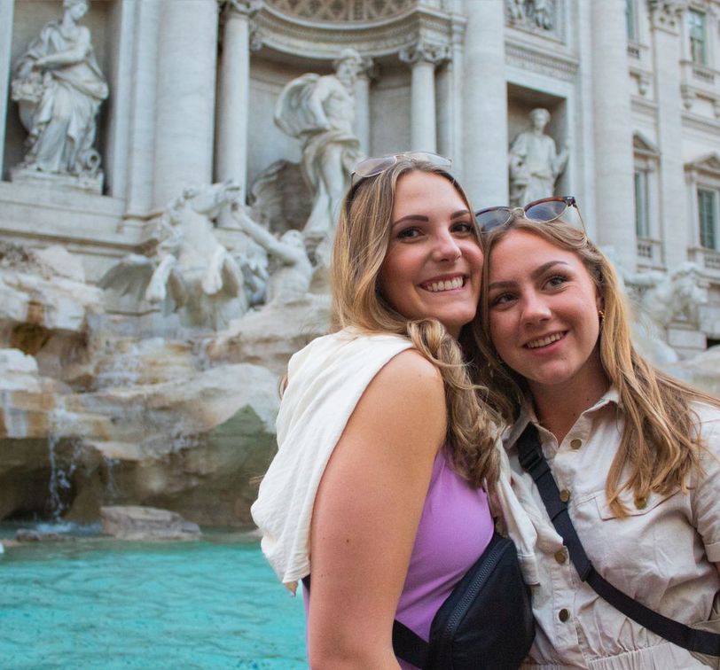 Two smiling women in front of the Trevi Fountain in Rome, with clear turquoise water and detailed marble sculptures in the background.