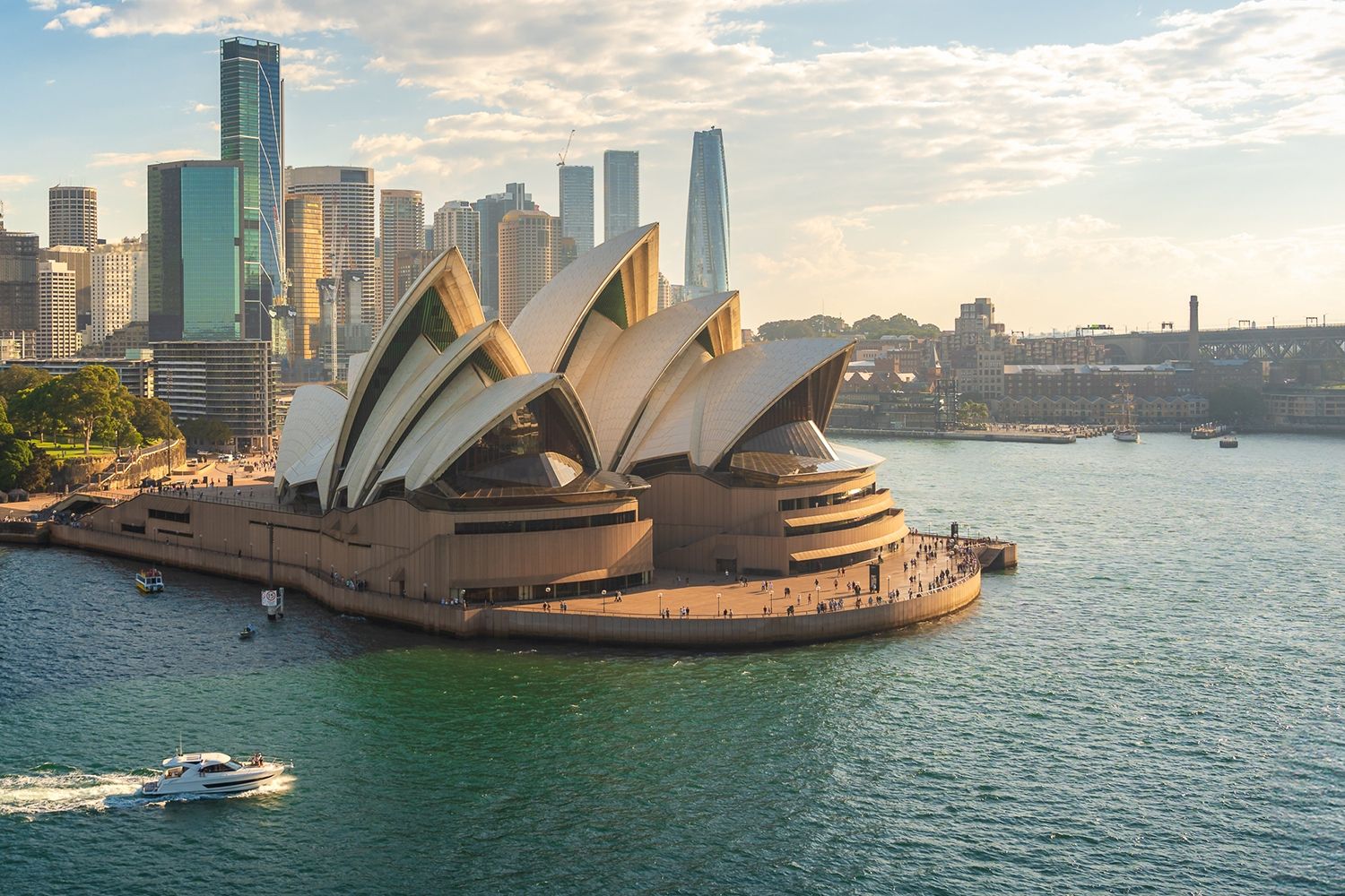 Sydney Opera House with its iconic sails by the waterfront, surrounded by skyscrapers, as a boat cruises in the harbor under a partly cloudy sky.