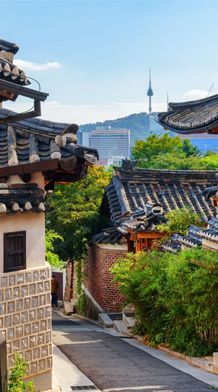 Traditional Korean hanok village with stone and wood houses, tile roofs, and a cityscape view in the distance under a clear blue sky.