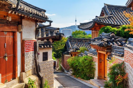 Traditional Korean hanok village with stone and wood houses, tile roofs, and a cityscape view in the distance under a clear blue sky.