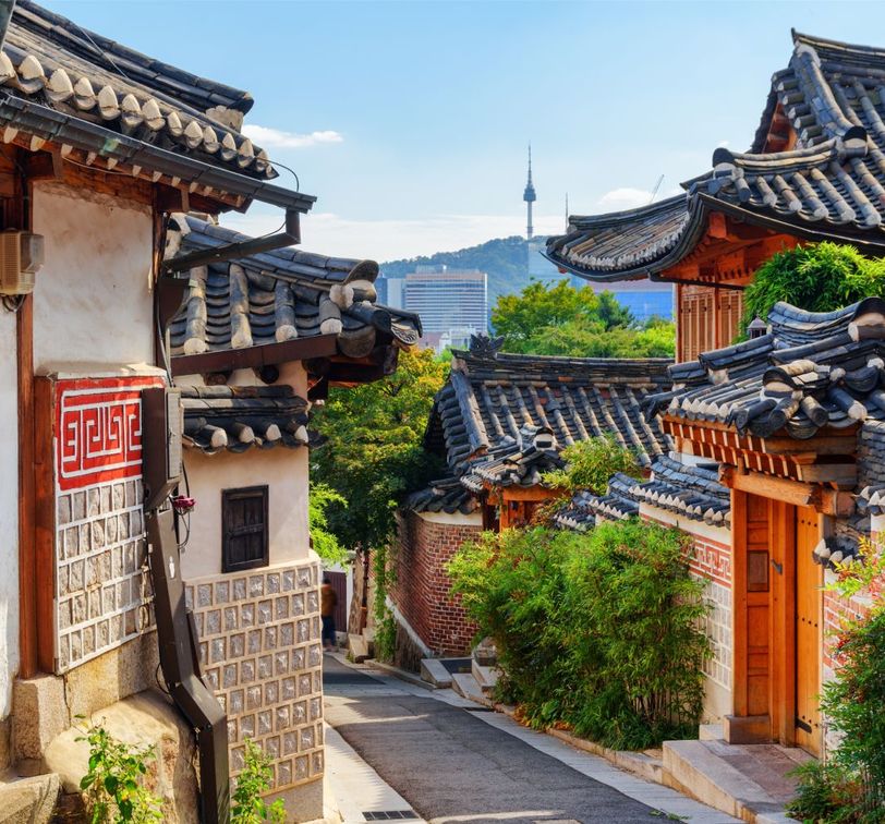 Traditional Korean hanok village with stone and wood houses, tile roofs, and a cityscape view in the distance under a clear blue sky.