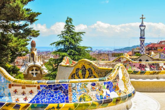Colorful mosaic benches in Park Güell, Barcelona, with a view of the city, lush greenery, and a tiled tower under a blue sky.