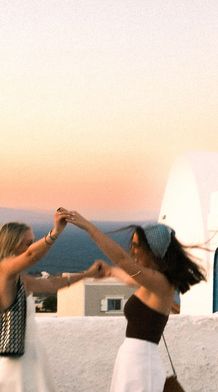 Two women dance joyfully on a rooftop at sunset, with a Greek-style building and the ocean in the background.