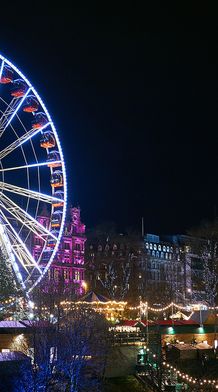 Illuminated Ferris wheel with colorful lights at a festive night scene, surrounded by buildings and trees adorned with string lights.