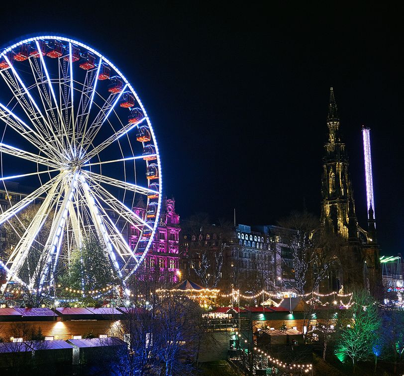 Illuminated Ferris wheel with colorful lights at a festive night scene, surrounded by buildings and trees adorned with string lights.