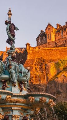A historic castle on a rocky hilltop bathed in golden sunlight, with an ornate fountain in the foreground surrounded by trees.