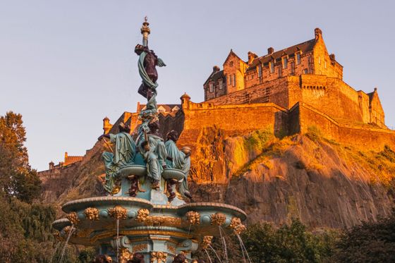 A historic castle on a rocky hilltop bathed in golden sunlight, with an ornate fountain in the foreground surrounded by trees.
