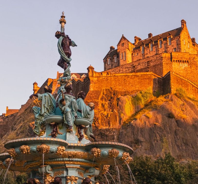 A historic castle on a rocky hilltop bathed in golden sunlight, with an ornate fountain in the foreground surrounded by trees.