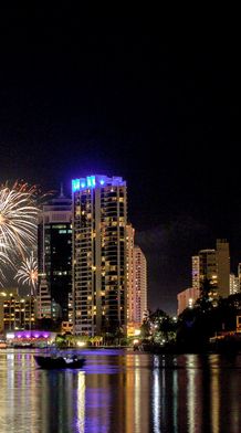 City skyline at night with illuminated skyscrapers and vibrant fireworks in the sky, reflecting on a calm body of water.