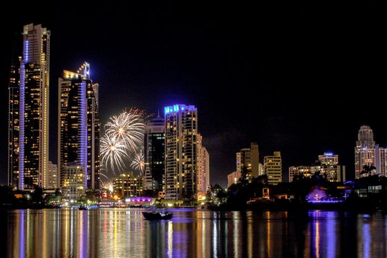 City skyline at night with illuminated skyscrapers and vibrant fireworks in the sky, reflecting on a calm body of water.