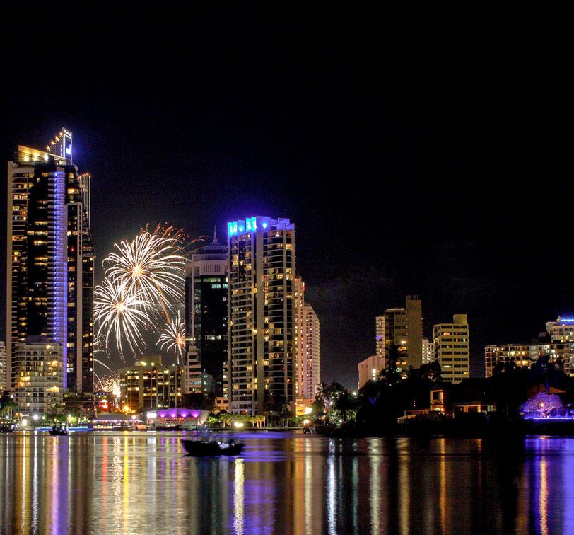 City skyline at night with illuminated skyscrapers and vibrant fireworks in the sky, reflecting on a calm body of water.