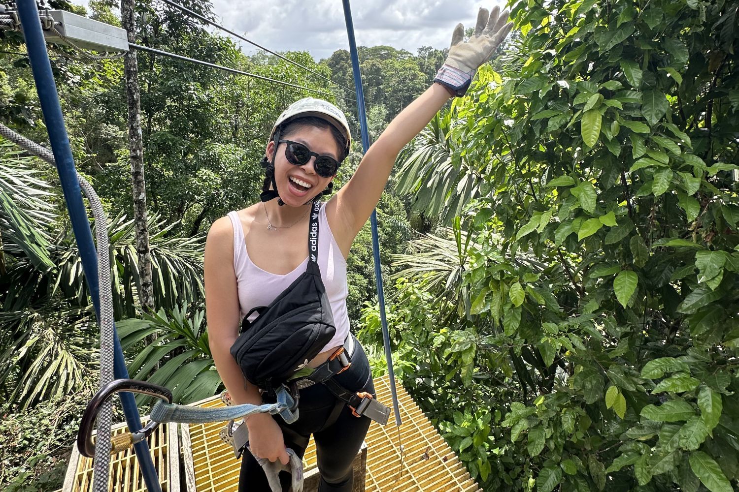 A woman wearing a helmet and sunglasses waves excitedly while on a zipline platform surrounded by lush green foliage.