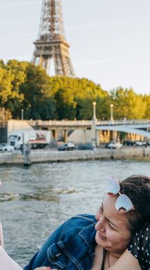 Three women smiling and laughing near a river, with the Eiffel Tower and greenery in the background on a sunny day.
