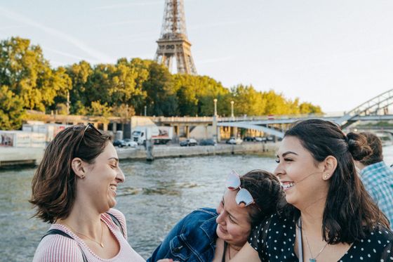 Three women smiling and laughing near a river, with the Eiffel Tower and greenery in the background on a sunny day.