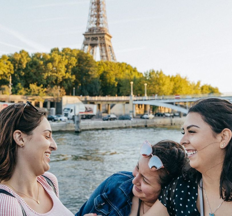 Three women smiling and laughing near a river, with the Eiffel Tower and greenery in the background on a sunny day.