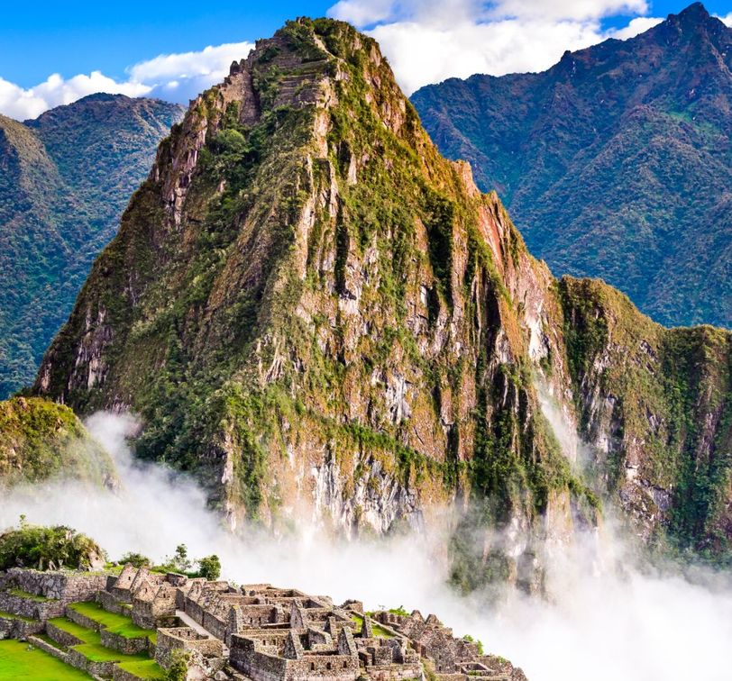 Ancient stone ruins of Machu Picchu surrounded by lush mountains and mist under a blue sky.