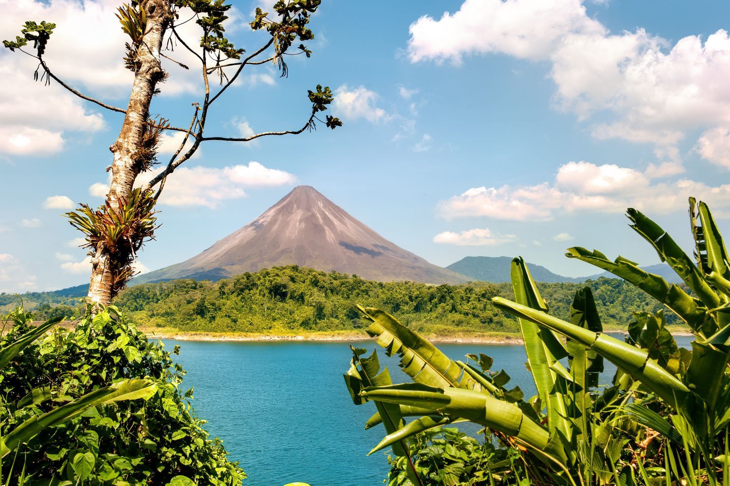 A picturesque volcano rising above a lush green forest, with a clear blue lake in the foreground and a few scattered clouds in the sky.
