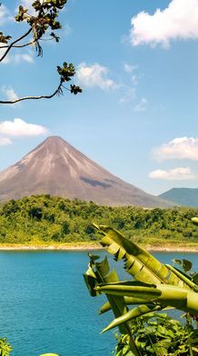 A picturesque volcano rising above a lush green forest, with a clear blue lake in the foreground and a few scattered clouds in the sky.