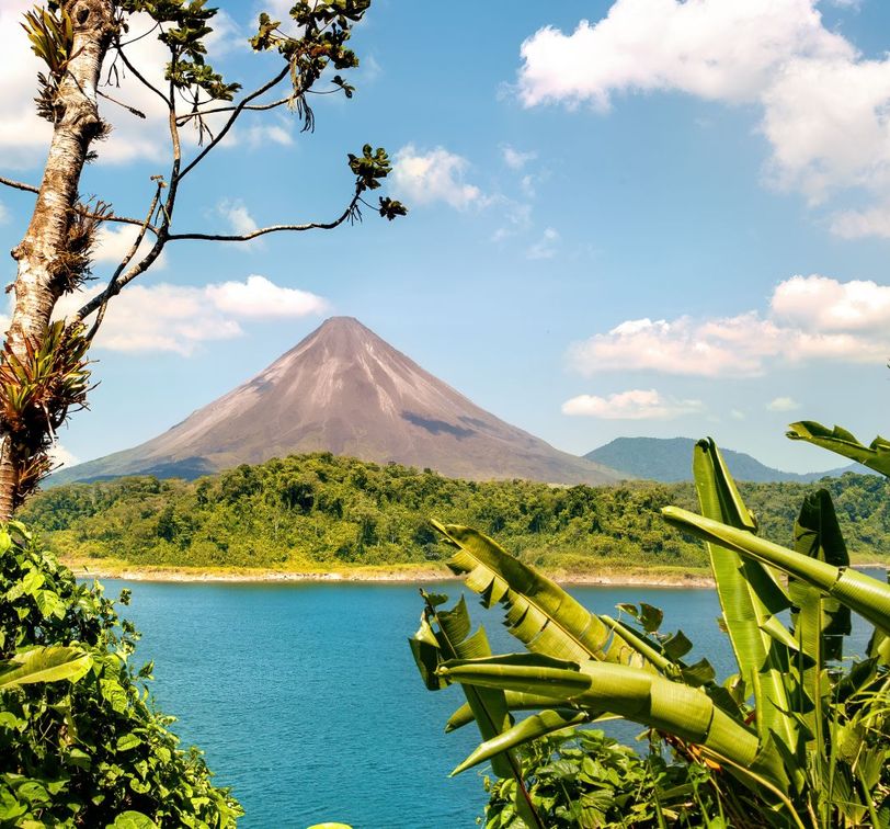 A picturesque volcano rising above a lush green forest, with a clear blue lake in the foreground and a few scattered clouds in the sky.