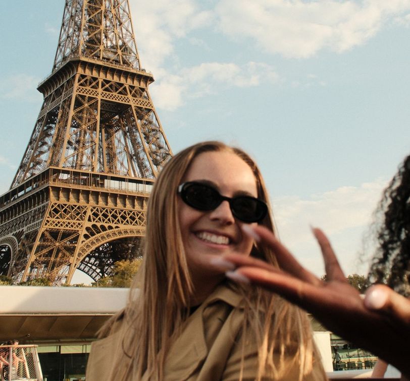 Two women smiling and wearing sunglasses with the Eiffel Tower in the background under a blue sky.