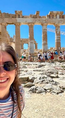 A person in sunglasses, holding a camera, smiles in front of the Parthenon with tourists in the background under a clear blue sky.