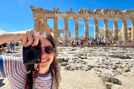 A person in sunglasses, holding a camera, smiles in front of the Parthenon with tourists in the background under a clear blue sky.