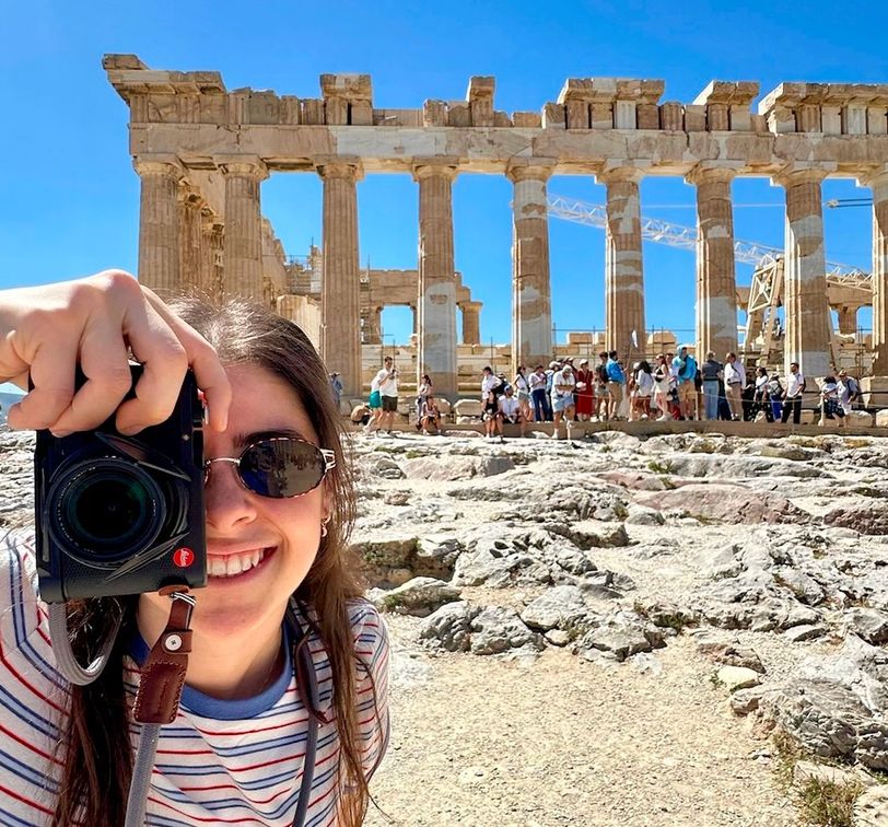A person in sunglasses, holding a camera, smiles in front of the Parthenon with tourists in the background under a clear blue sky.