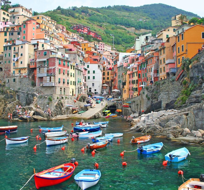 Colorful houses on a steep hillside overlook a small harbor with numerous boats floating on clear turquoise waters.