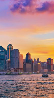 Hong Kong skyline at sunset, with tall skyscrapers, a ferris wheel, and boats on Victoria Harbour under a colorful sky.