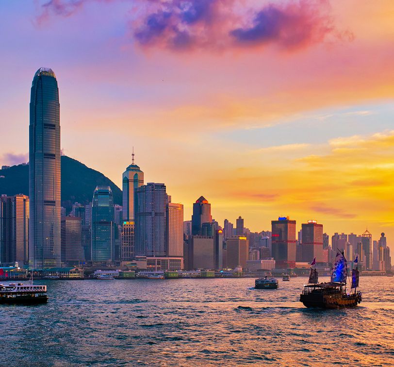 Hong Kong skyline at sunset, with tall skyscrapers, a ferris wheel, and boats on Victoria Harbour under a colorful sky.
