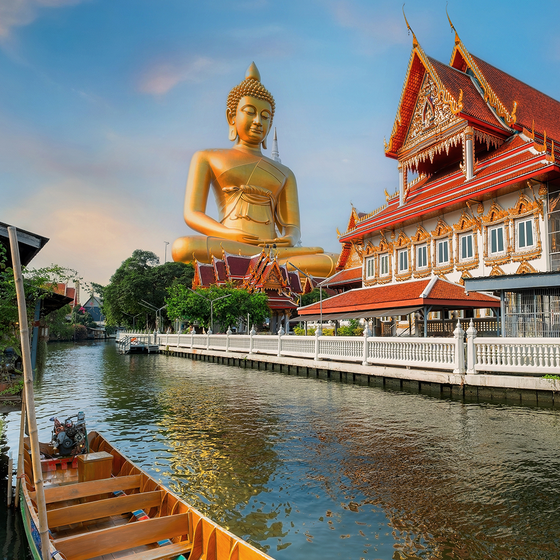A large golden Buddha statue overlooks a canal beside a traditional Thai temple with ornate red roofs and gold detailing under a blue sky.