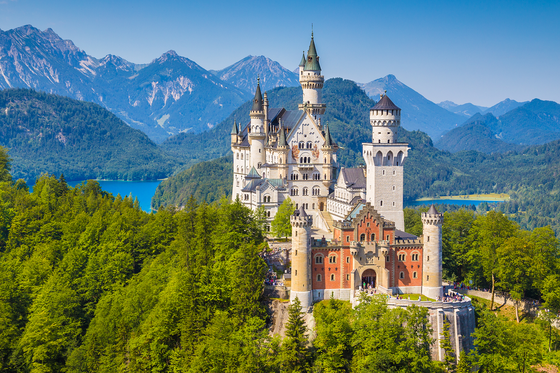 A fairy tale castle with multiple turrets and towers, nestled amidst lush green forests and mountains under a clear blue sky.