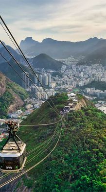 Aerial view of Rio de Janeiro with a cable car, lush mountains, cityscape, and coastline under a cloudy sky.