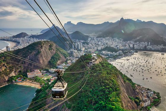Aerial view of Rio de Janeiro with a cable car, lush mountains, cityscape, and coastline under a cloudy sky.
