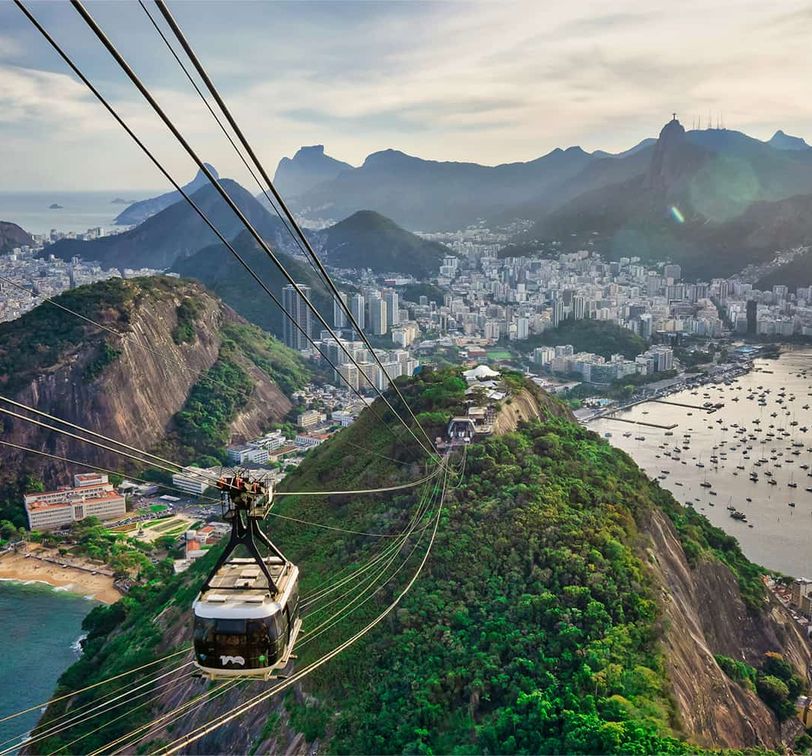 Aerial view of Rio de Janeiro with a cable car, lush mountains, cityscape, and coastline under a cloudy sky.