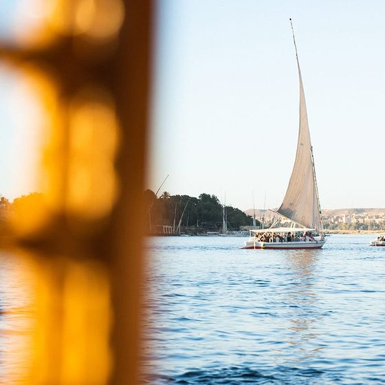 Sailboats with tall white sails on a calm river, framed by a blurred foreground, with distant hills and clear skies in the background.