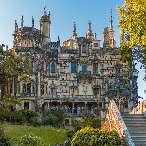 Ornate Gothic Revival-style palace with intricate details, set amidst lush greenery, with a stone staircase leading up to the entrance.